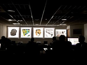 In a dark room, people sit in front of a wall where five images are being projected.