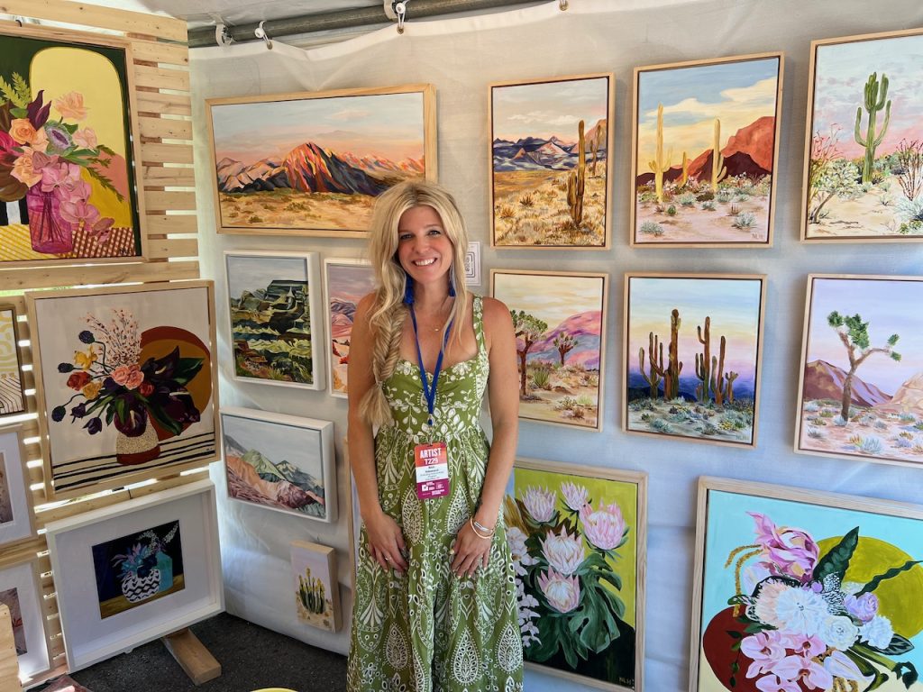 Female artist smiling and standing in front of a display of several paintings.