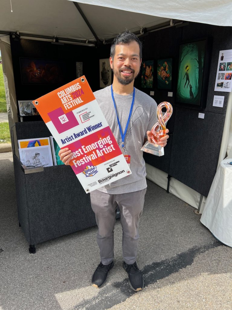 A male artist smiling and holding a trophy and a poster in front of his art booth.